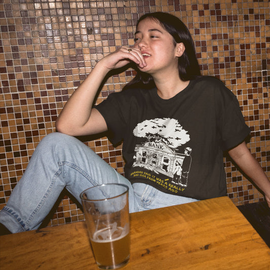 Person sitting at a table with a glass of beer, wearing a black t-shirt with a graphic design. A Banksy style stencil art t-shirt with an anti-capitalism message. A thought provoking future where money and banking are a dark memory and our children have something more natural to look forward to. Made from 100% organic ring-spun cotton, this utopian graffiti stencil t-shirt is unisex, high-quality, super comfy, and best of all, eco-friendly.