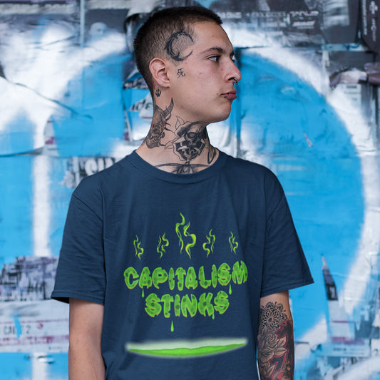 A tattooed man standing against a graffiti wall wearing a French navy blue short sleeved crew neck t-shirt with "CAPITALISM STINKS" printed in a mouldy green cheese style.