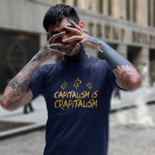 A tattooed man standing in the street with his hands over his face, wearing a French navy blue short sleeved crew neck t-shirt With the graphic text saying "CAPITALISM IS CRAPITALISM" with whiffs of vapor above it.