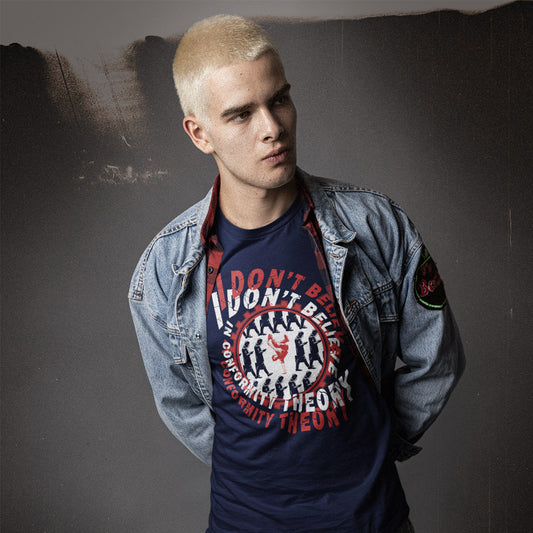 A young man with short blond hair wears a navy graphic t-shirt under an open light-wash denim jacket. The t-shirt features a red and white circular graphic with the text 'I DON'T BELIEVE IN CONFORMITY THEORY'.
