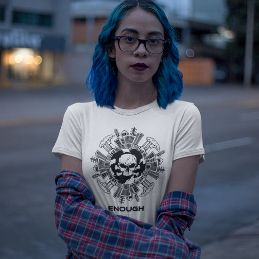 A person with bright blue hair and glasses stands outdoors wearing a cream graphic T-shirt with a black skull-and-tools print and the word 'ENOUGH' below, along with a red-and-blue plaid shirt draped off the shoulders.