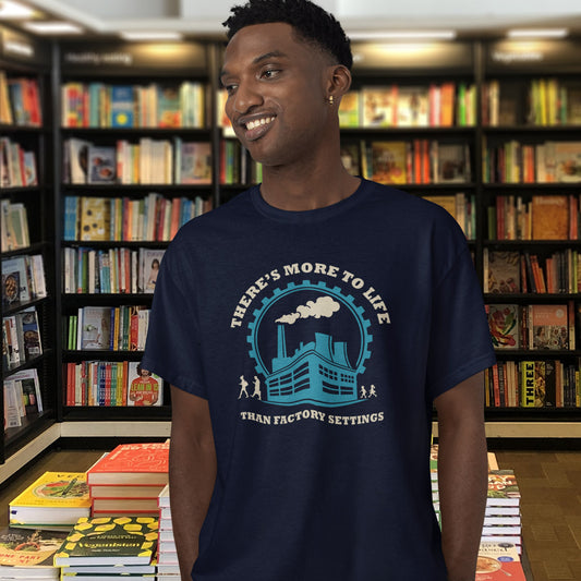 A man wearing a French navy blue graphic t-shirt in a bookshop. The t-shirt features a light blue and white graphic of a factory with the text 'THERE'S MORE TO LIFE THAN FACTORY SETTINGS'.
