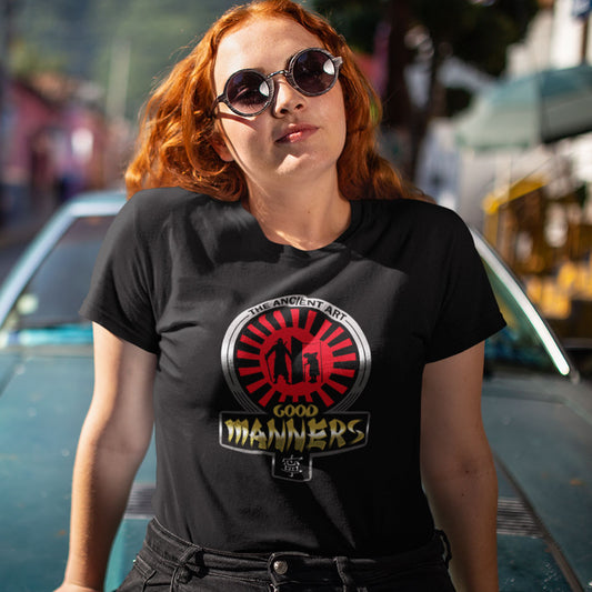 A woman with red hair and sunglasses wearing a black graphic t-shirt with a 'The Ancient Art Good Manners' emblem, posed outdoors in front of a blurred car.