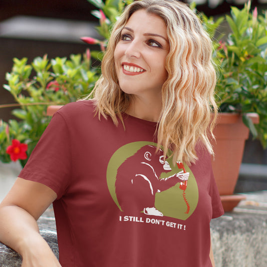 Woman wearing a burgundy t-shirt with a graphic design and text, standing outdoors with plants in the background. This satirical Banksy style stencil art t-shirt design highlights the human obsession with smart phones and how strange it must look to an ape. An outsiders view of a world glued to mobile phones. Made from 100% organic ring-spun cotton, this street art social awareness t-shirt is unisex, high-quality, super comfy, and best of all, eco-friendly.