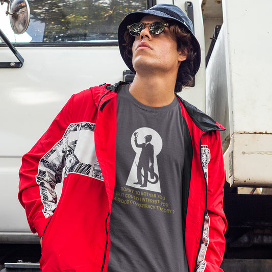 A man wearing a red hooded jacket with black and white graphic panels and an anthracite grey graphic t-shirt with a white graphic features a white keyhole silhouette of a reptilian salesman holding his hat, with gold text underneath reading 'SORRY TO BOTHER YOU BUT COULD I INTEREST YOU IN A RELIABLE CONSPIRACY THEORY?'.