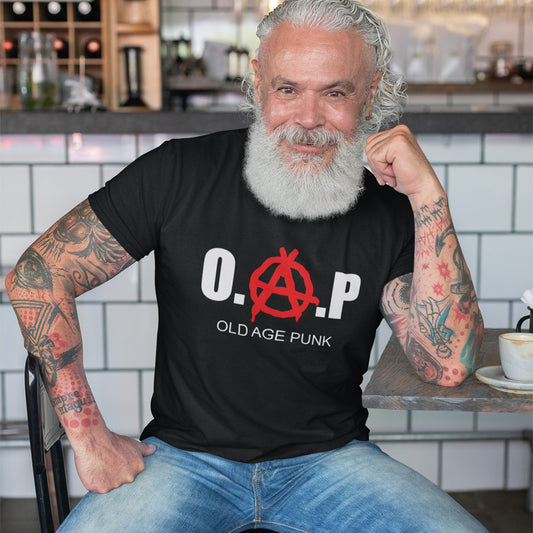A grey haired, bearded man sitting at a table, smiling and wearing a black short sleeved crew neck t-shirt with the text graphic "PRO CHOICE, ANTI RACIST, PLANT BASED, ANIMAL LOVER" in black.