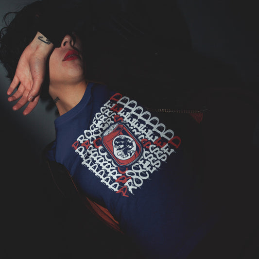 A woman with her arm over her face, in a dark room wearing a French navy blue crew neck t-shirt with a graphic of a baby in a soda can on the phone, surrounded by the text "PROCESSED" in white and red.