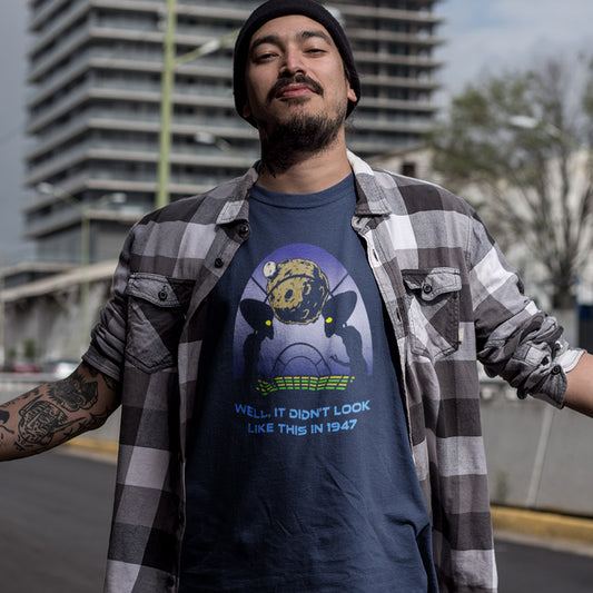 A man wearing a navy blue graphic t-shirt under an open black and white plaid flannel shirt, with a black beanie. The t-shirt features a graphic of a skull wearing a space helmet with the text 'WELL IT DIDN'T LOOK LIKE THIS IN 1947'.