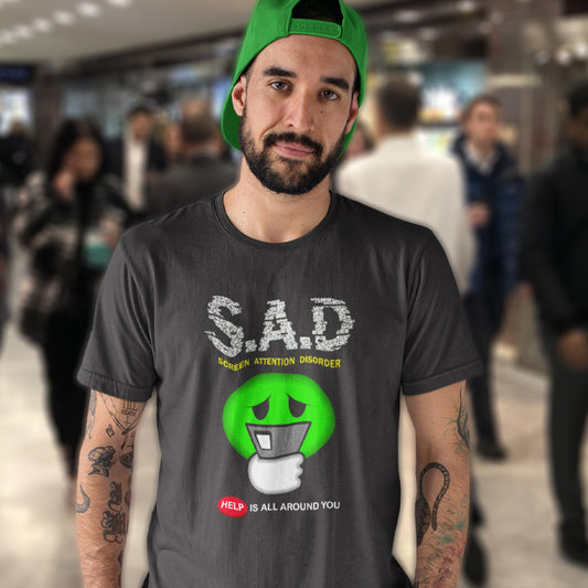 A man in a shopping centre wearing a green cap and an anthracite grey t-shirt with " S.A.D Screen attention disorder" text and a sick looking emoji face.