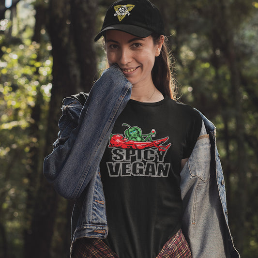 A woman wearing a black t-shirt with a 'Spicy Vegan' graphic featuring a graphic print of a green chili pepper and a red chilli character with the text 'SPICY VEGAN' underneath., styled with a denim jacket and a black cap in a forest setting.