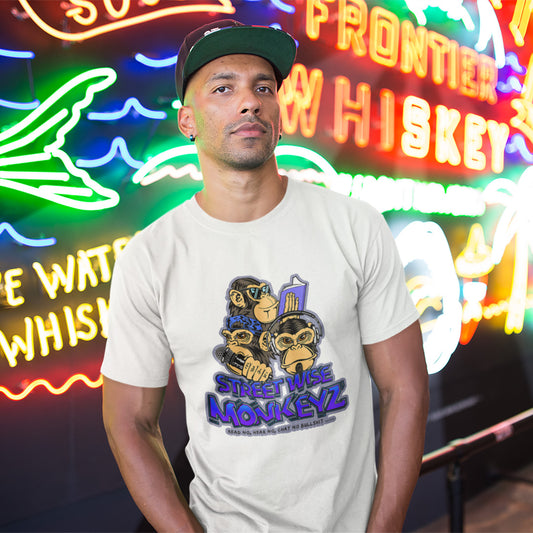 A man in a cap standing in front of neon lights, wearing a white short-sleeve crewneck t-shirt featuring a colourful graphic print of three cartoon monkeys with the text 'STREET WISE MONKEYZ' in purple and blue.