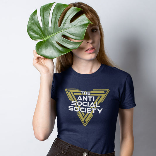 A woman wearing a French navy blue t-shirt with a gold and white graphic that reads 'THE ANTI SOCIAL SOCIETY' holds a large green monstera leaf in front of her face, set against a plain light grey background.