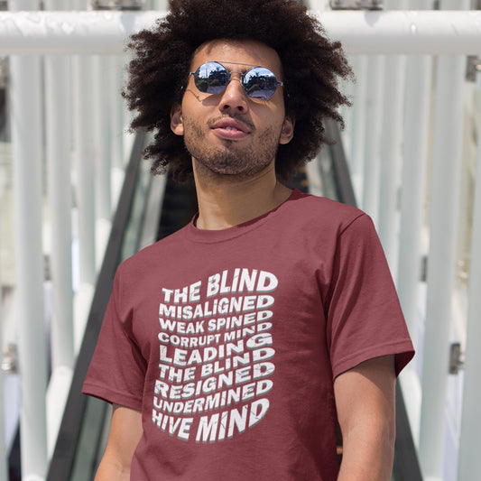 A man standing on a bridge, wearing sunglasses and a burgundy t-shirt with a white graphic text print on the front reading 'THE BLIND MISALIGNED WEAK SPINED CORRUPT MIND LEADING THE BLIND RESIGNED UNDERMINED HIVE MIND' in a stacked, stylized font.