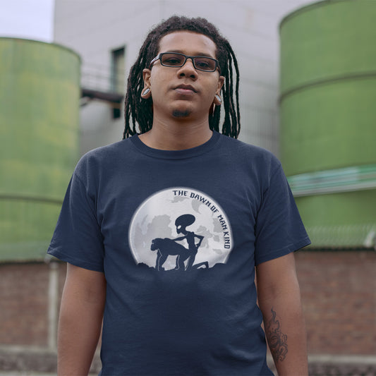A man with dreadlocks standing in front of water barrels, wearing a French navy blue, short sleeved, crew neck t-shirt with the silhouette of an alien and a monkey in front of the moon with the text "THE DAWN OF HUMANKIND".