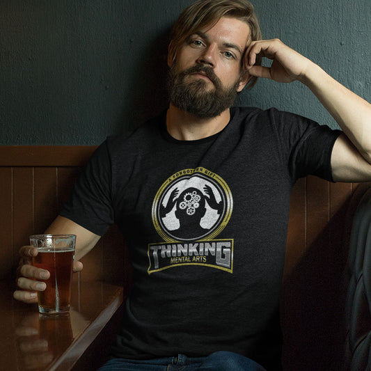 A man with a beard sits in a dimly lit booth wearing a black graphic t-shirt with a circular 'Thinking Mental Arts' logo. He holds a glass of amber beer.