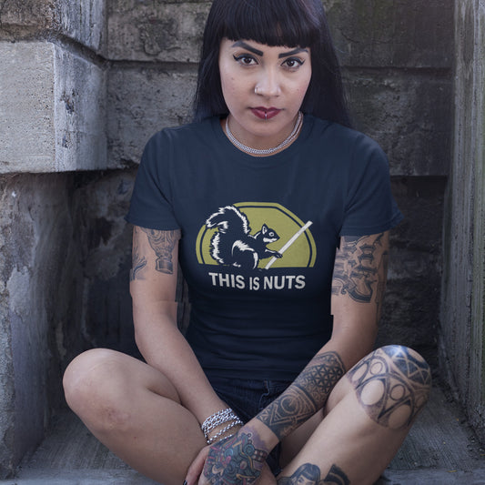 A woman with black hair and tattoos, sitting on a step wearing a French navy blue t-shirt featuring a graphic of a squirrel holding a smartphone in front of a yellow-green circle, with the text 'THIS IS NUTS' printed below in white.