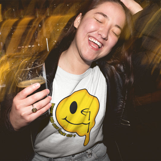 A laughing woman with her hand in her hair, holding a beer and wearing a white crew neck t-shirt with a yellow dripping smiley face and the text "WHERE DID OUR LOVE GO?".