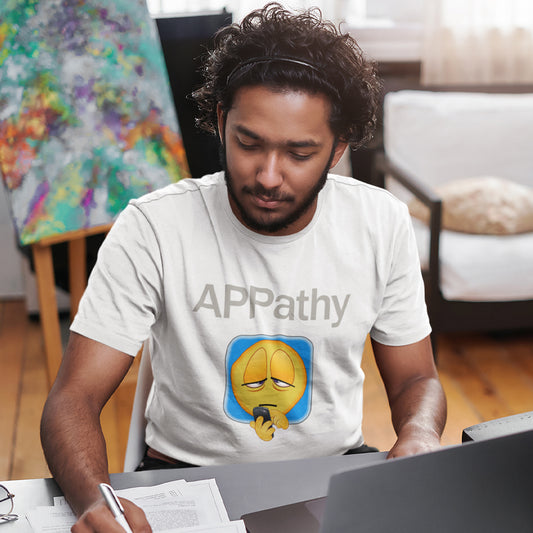 Man wearing a white t-shirt with an emoji design and 'APPathy' text, sitting at a desk. This anti mobile phone abuse t shirt promotes social awareness of the amount of apps we now need in modern society. This unique emoji t-shirt design is for everyone who doesn't feel they are a child of the mobile phone. Made from 100% organic ring-spun cotton, this anti smart phone abuse t shirt is unisex, high-quality, super comfy, and best of all, eco-friendly.
