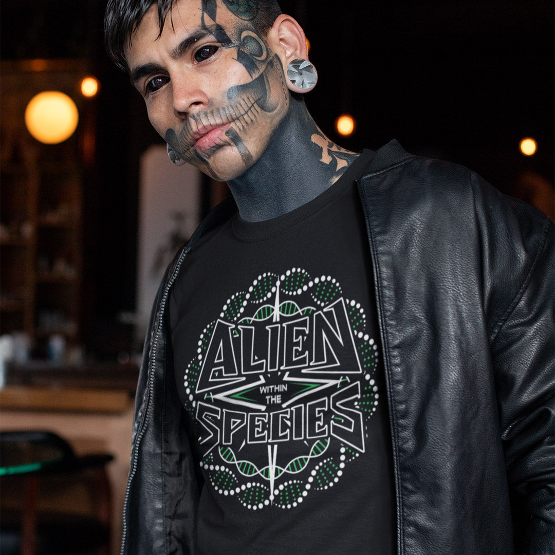 Man wearing a black t-shirt with 'Alien Within the Species' design, standing in a dimly lit room. This non conformist t shirt expands the use of the word 'Alien'. This design is ideal for everyone that feels they don't truly relate to the rest of humankind and modern society. Made from 100% organic ring-spun cotton, this counter culture t-shirt is unisex, high-quality, super comfy, and best of all, eco-friendly.