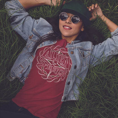 Person wearing a red t-shirt with 'Alien within the species' design, denim jacket, and sunglasses, lying on grass. This non conformist t shirt expands the use of the word 'Alien'. This design is ideal for everyone that feels they don't truly relate to the rest of humankind and modern society. Made from 100% organic ring-spun cotton, this counter culture t-shirt is unisex, high-quality, super comfy, and best of all, eco-friendly.