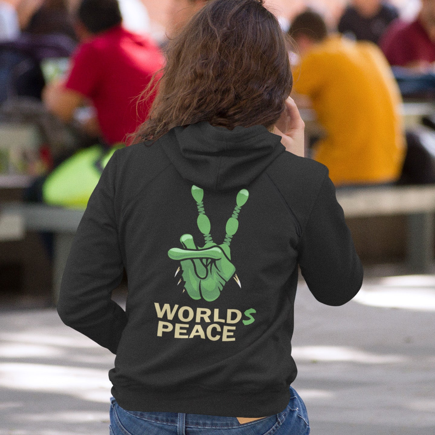 Person wearing a black hoodie with green hand symbol and 'Worlds Peace' text, standing in a casual setting. A world peace hoodie design with an alien lovers twist. When we think of world peace lets not be selfish about it., lets include all Worlds. Comfortable and practical. It's extra soft and has a convenient front pouch pocket. Made from organic cotton and recycled polyester, this funny alien hoodie is the perfect eco-friendly choice.