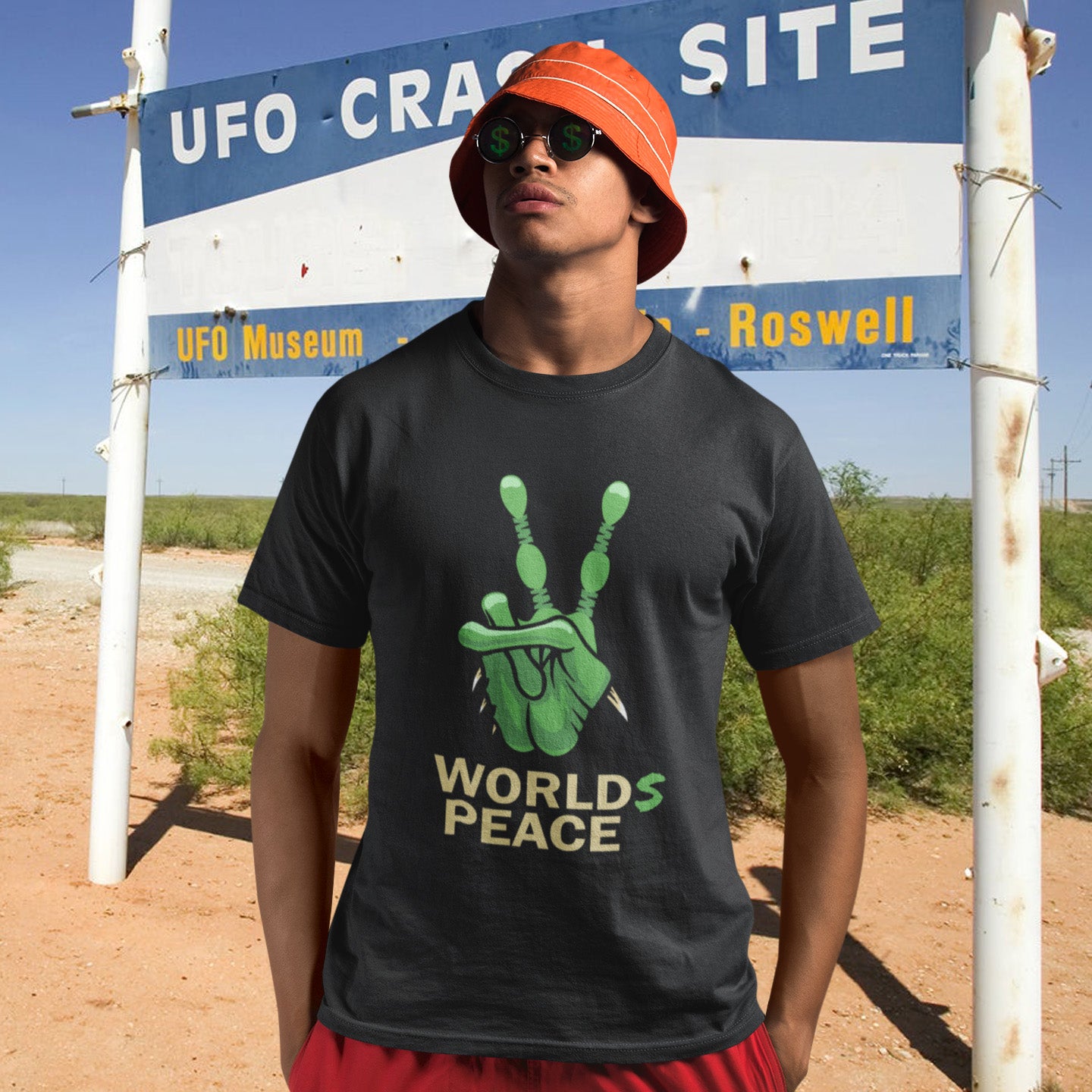 Person wearing a black t-shirt with green graphic and text, standing in front of a UFO crash site sign in Roswell. A world peace t shirt with an alien lovers twist. When we think of world peace lets not be selfish about it., lets include all Worlds. Made from 100% organic ring-spun cotton, this funny alien t shirt is unisex, high-quality, super comfy, and best of all, eco-friendly.