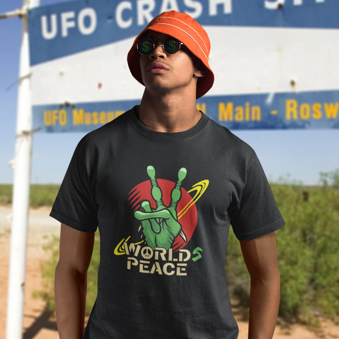 A man wearing a black graphic t-shirt with a green alien hand making the peace sign over a red planet graphic and the words 'WORLD'S PEACE'. He is also wearing an orange bucket hat and sunglasses, standing in front of a 'UFO CRASH SITE' sign in a desert landscape.