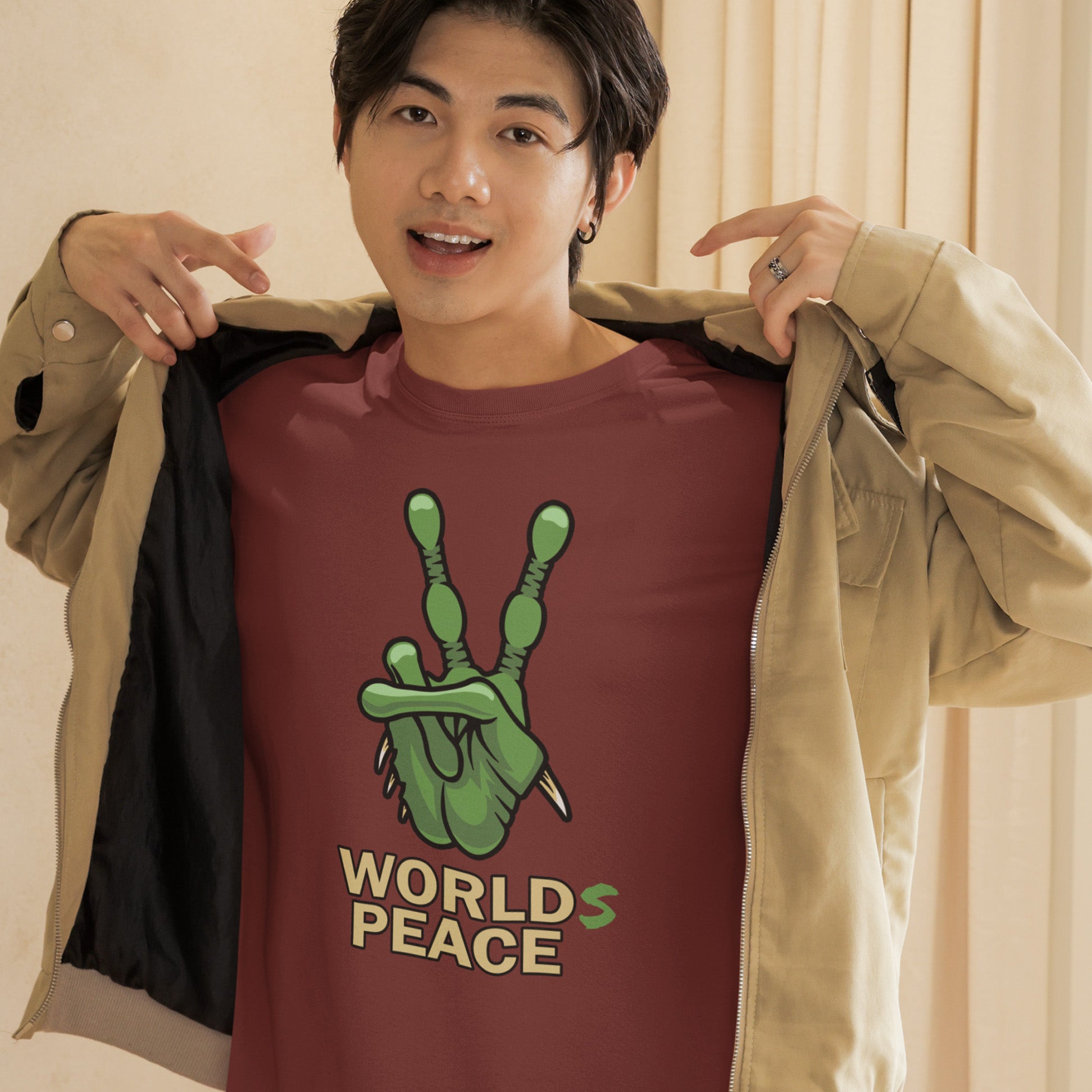 Person wearing a maroon t-shirt with a green graphic and 'World's Peace' text, holding a beige jacket. A world peace t-shirt with an alien lovers twist. When we think of world peace lets not be selfish about it., lets include all Worlds. Made from 100% organic ring-spun cotton, this humorous anti-war alien t-shirt is unisex, high-quality, super comfy, and best of all, eco-friendly.