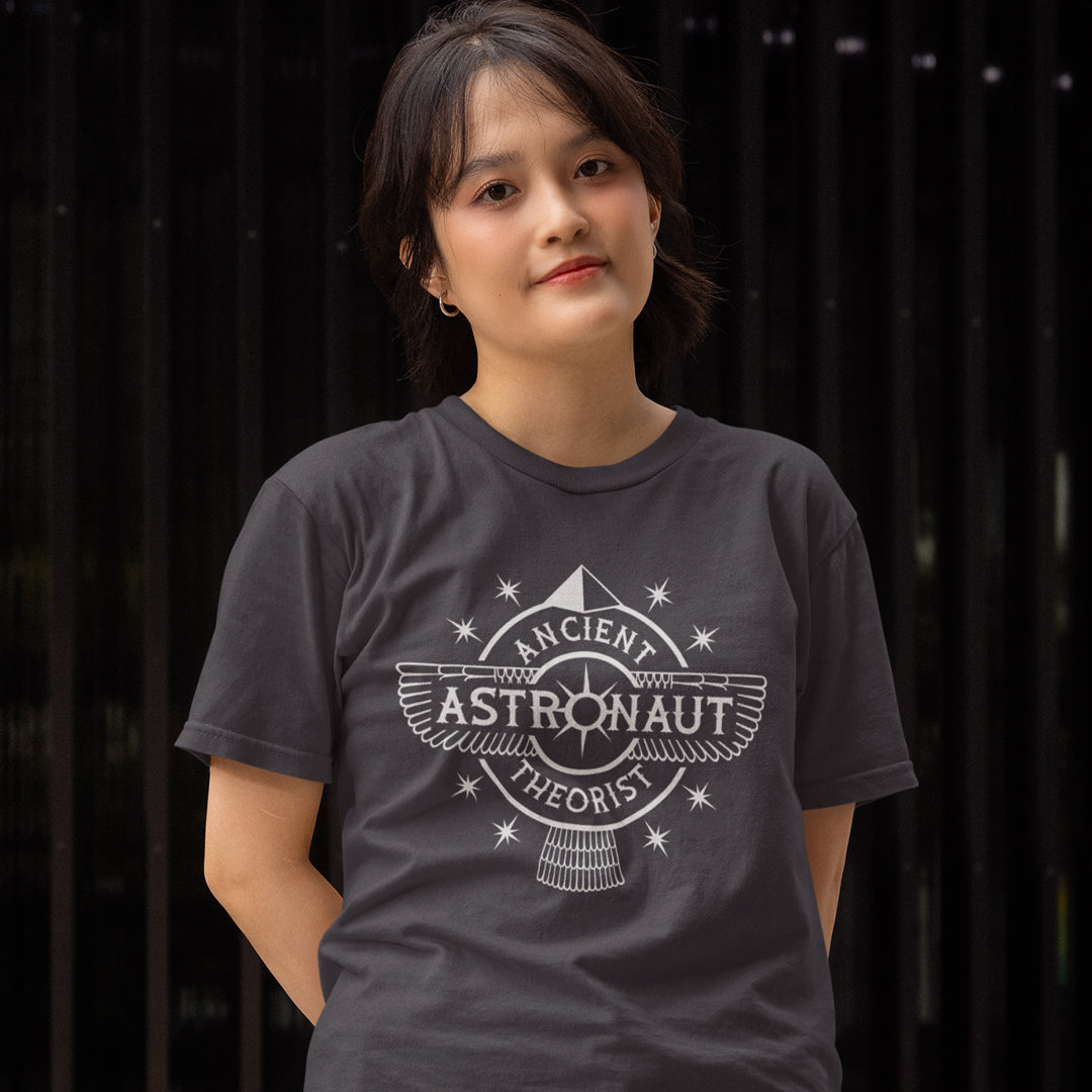 Woman standing with her hands behind her back earing an anthracite grey t-shirt  with "ANCIENT ASTRONAUT THEORIST" logo.