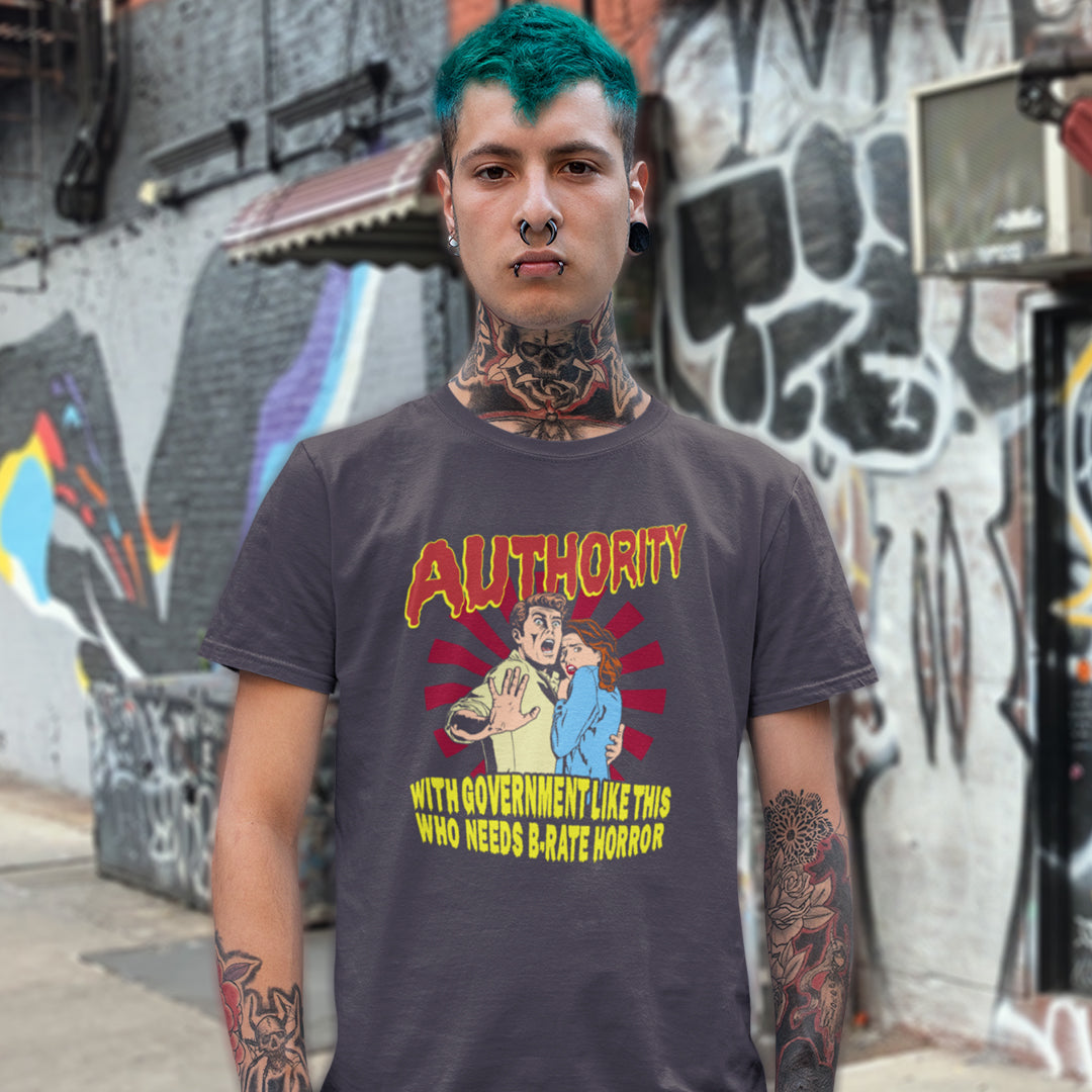 A man with green hair, tattoos, and a nose ring stands in front of a graffiti-covered wall wearing a dark gray graphic t-shirt with a red and yellow 'AUTHORITY' print.