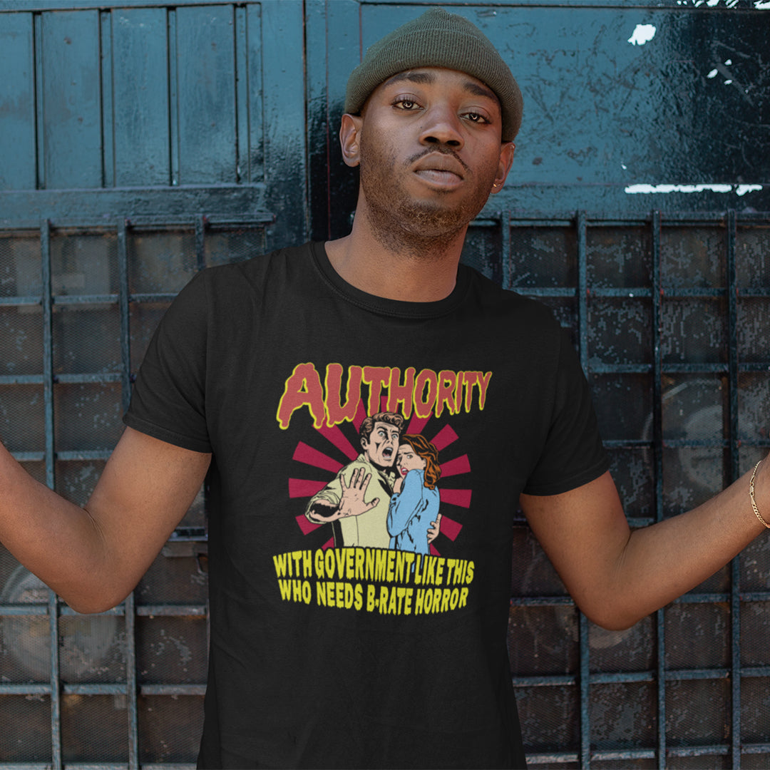 A man wearing a black graphic t-shirt and a green beanie stands with arms outstretched in front of a dark blue metal gate. The t-shirt features a retro-style horror graphic with the word 'AUTHORITY' in red and yellow, and a cartoon illustration of a man and a blue monster, with the text 'WITH GOVERNMENT LIKE THIS WHO NEEDS B-RATE HORROR' below.