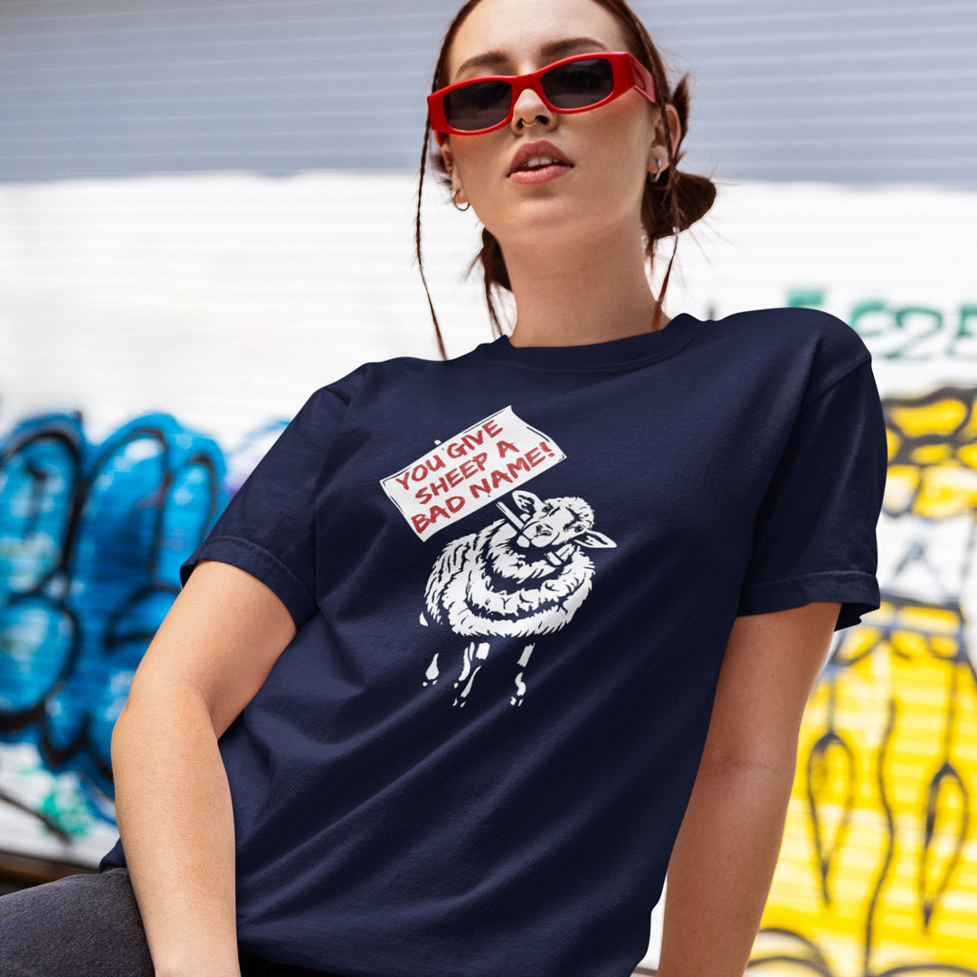 A woman wearing a French navy blue graphic t-shirt with a white sheep illustration and a sign that reads 'YOU GIVE SHEEP A BAD NAME!'. She is wearing red sunglasses and posing in front of a graffiti-covered wall.