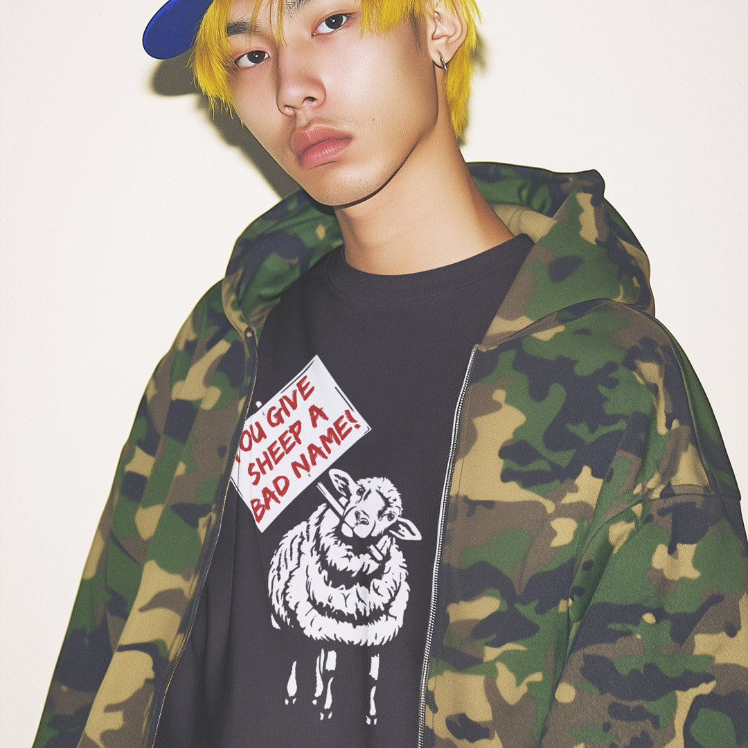A model wearing an anthracite grey graphic t-shirt with a white sheep print and a green camouflage zip-up hoodie, paired with a blue cap, against a plain off-white background.