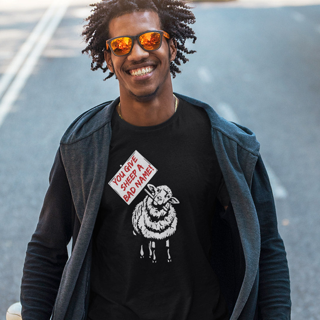 A smiling man wearing a black graphic t-shirt with a white sheep illustration and a red-and-white sign reading 'YOU GIVE SHEEP A BAD NAME!' stands outdoors on a paved road. He is also wearing a dark gray zip-up hoodie and orange-tinted sunglasses.