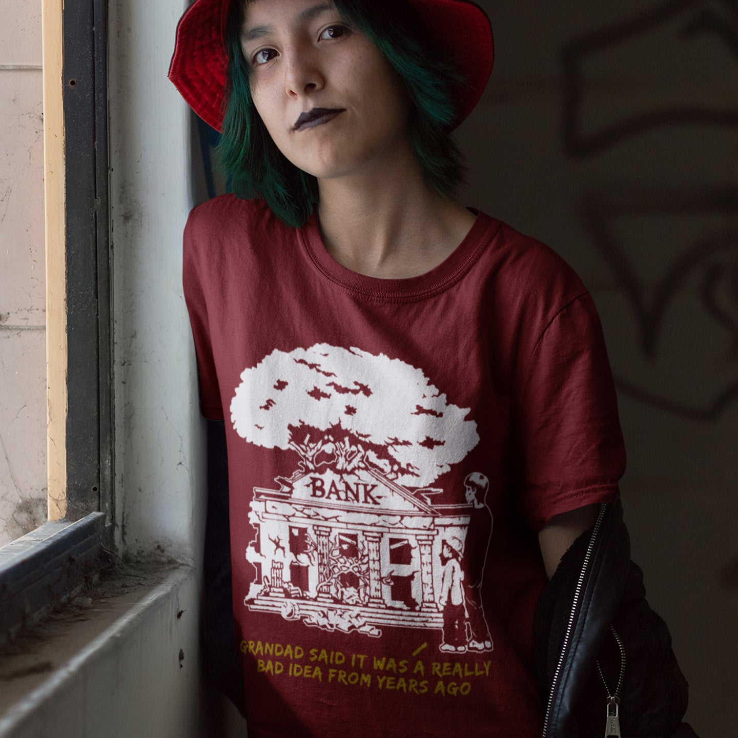 Person wearing a red t-shirt with a graphic design and text, standing indoors. A Banksy style stencil art t-shirt with an anti-capitalism message. A thought provoking future where money and banking are a dark memory and our children have something more natural to look forward to. Made from 100% organic ring-spun cotton, this utopian graffiti stencil t-shirt is unisex, high-quality, super comfy, and best of all, eco-friendly.