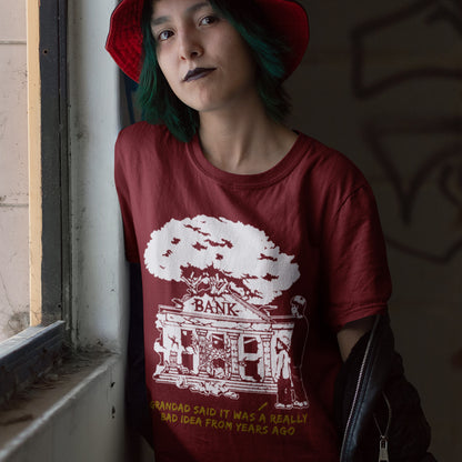 Person wearing a red t-shirt with a graphic design and text, standing indoors. A Banksy style stencil art t-shirt with an anti-capitalism message. A thought provoking future where money and banking are a dark memory and our children have something more natural to look forward to. Made from 100% organic ring-spun cotton, this utopian graffiti stencil t-shirt is unisex, high-quality, super comfy, and best of all, eco-friendly.