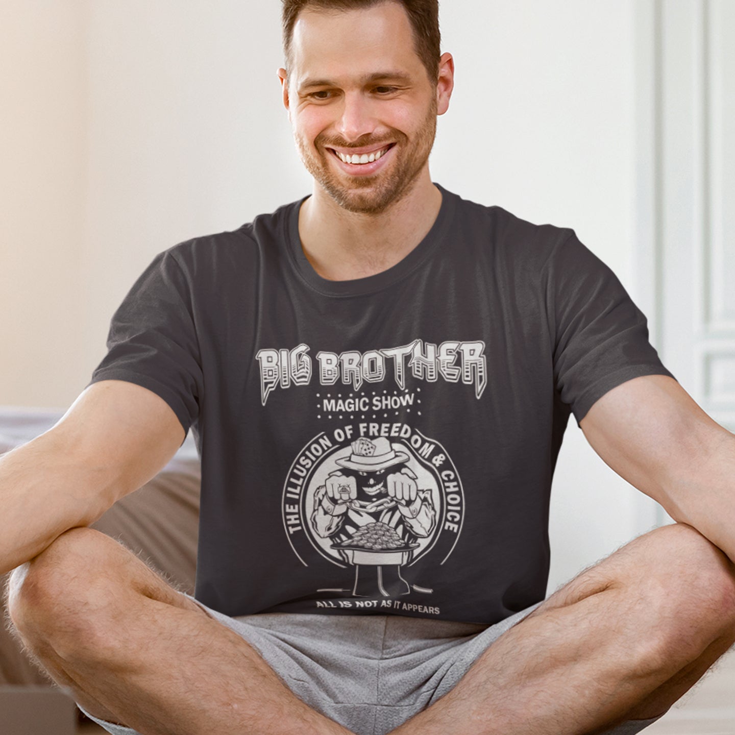 Man wearing a anthracite t-shirt with graphic design sitting on a couch. A funny conspiracy theory t-shirt suggesting our 'freedom of choice' is just part of an elaborate illusion. Magic shows are great until you realise you are living in one. This witty unique design hints at our society being distracted by greed. Made from 100% organic ring-spun cotton, this witty conspiracy theory t-shirt is unisex, high-quality, super comfy, and best of all, eco-friendly.