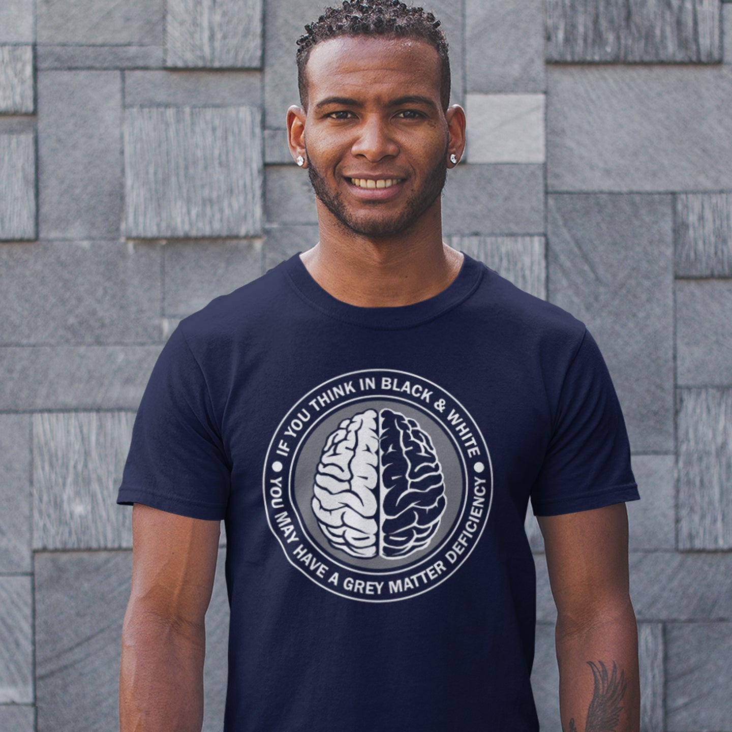 Man wearing a French navy blue t-shirt with a brain graphic and text against a stone wall background. A stencil art t shirt design. It would be so easy if life was black and white, but it's the grey area where all the thinking and personal growth is done. A philosophy t shirt design for people who see the detail and can think outside the box. Made from 100% organic ring-spun cotton, this social awareness t shirt is unisex, high-quality, super comfy, and best of all, eco-friendly.