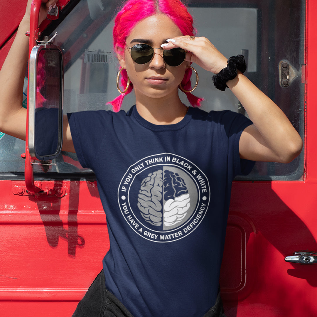 A woman with pink hair, standing against a van door, wearing sunglasses and a French navy blue short sleeved crew neck t-shirt with a graphic of a brain with the text" IF YOU ONLY THINK IN BLACK & WHITE, YOU HAVE A GREY MATTER DEFICIENCY" around it.
