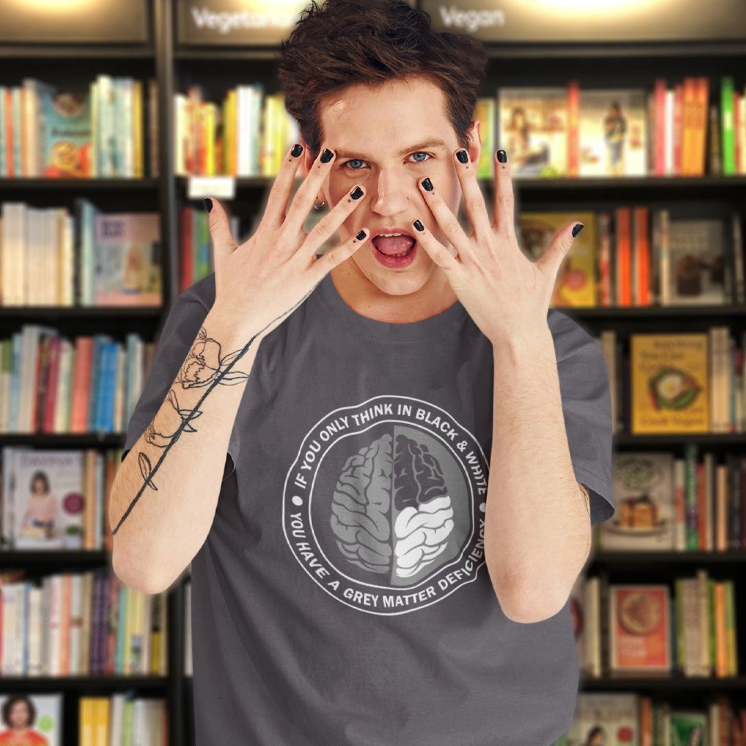 A man holding his hands up while standing in a book store, wearing black nail vanish and an anthracite grey short sleeved crew neck t-shirt with a graphic of a brain with the text" IF YOU ONLY THINK IN BLACK & WHITE, YOU HAVE A GREY MATTER DEFICIENCY" around it.
