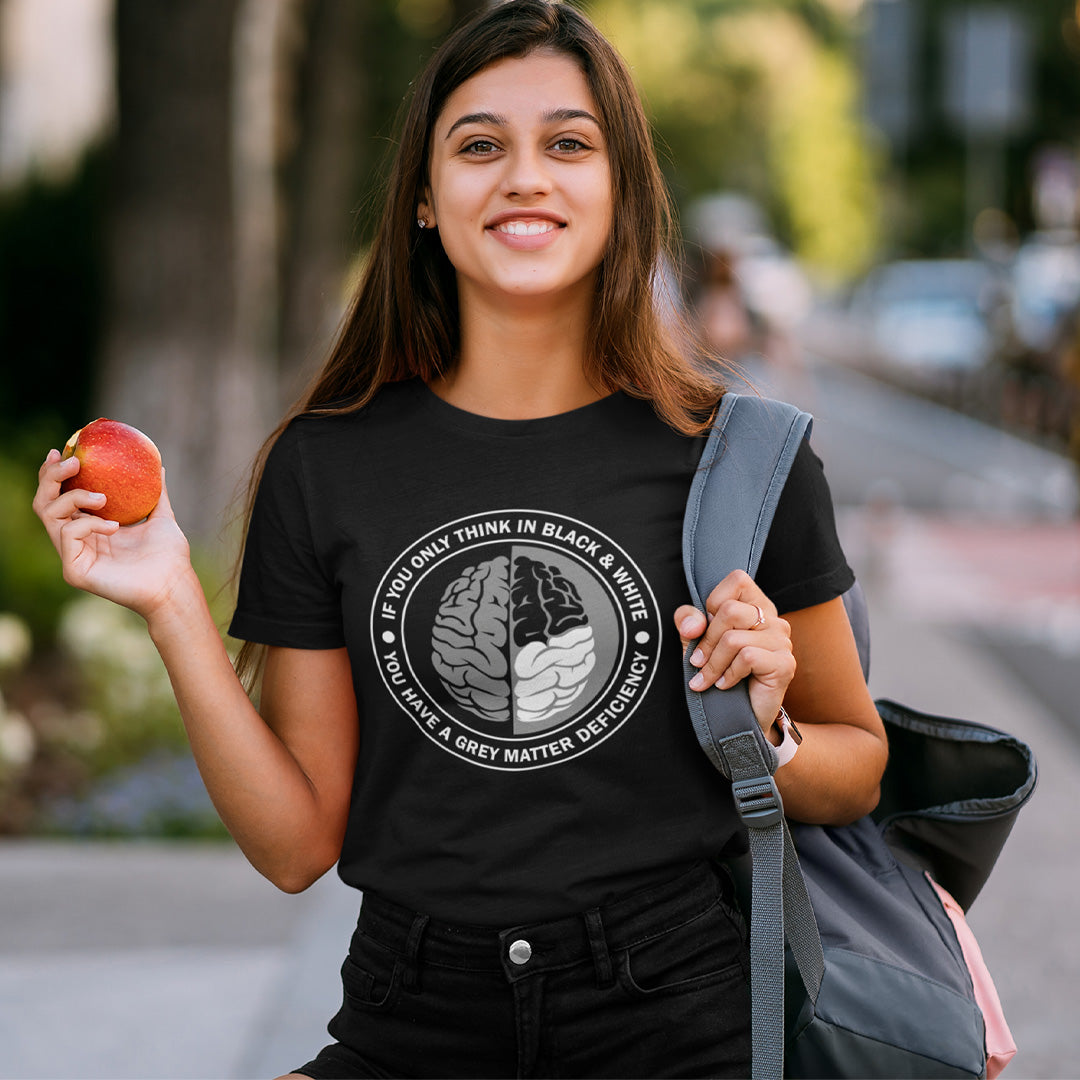 A woman with long brown hair, smiling and holding an apple, wearing a black short sleeved crew neck t-shirt with a graphic of a brain with the text" IF YOU ONLY THINK IN BLACK & WHITE, YOU HAVE A GREY MATTER DEFICIENCY" around it.