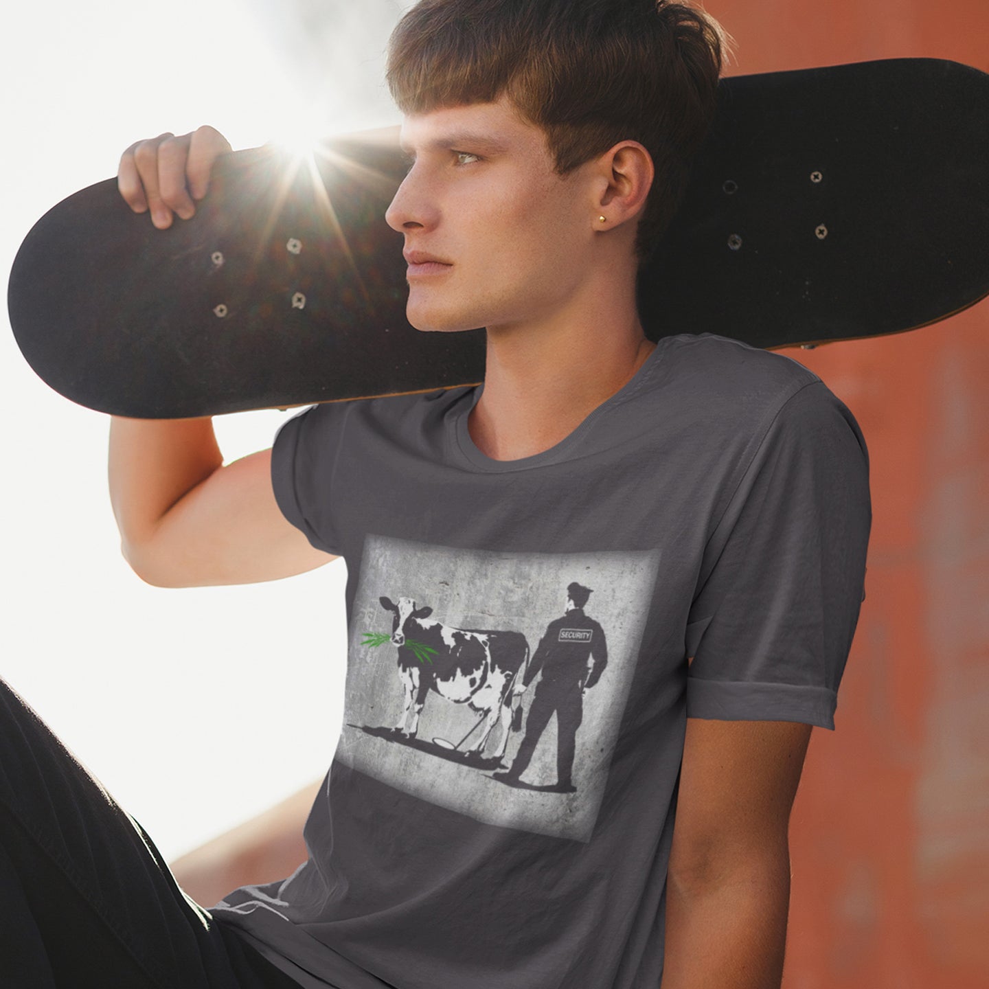 Person wearing a t-shirt with a graphic design of a cow and a person, holding a skateboard. A stencil graffiti t shirt showing the thin line between security and invasion of privacy, highlighted with a touch of satire and sarcasm. Made from 100% organic ring-spun cotton, this urban art t shirt is unisex, high-quality, super comfy, and best of all, eco-friendly.