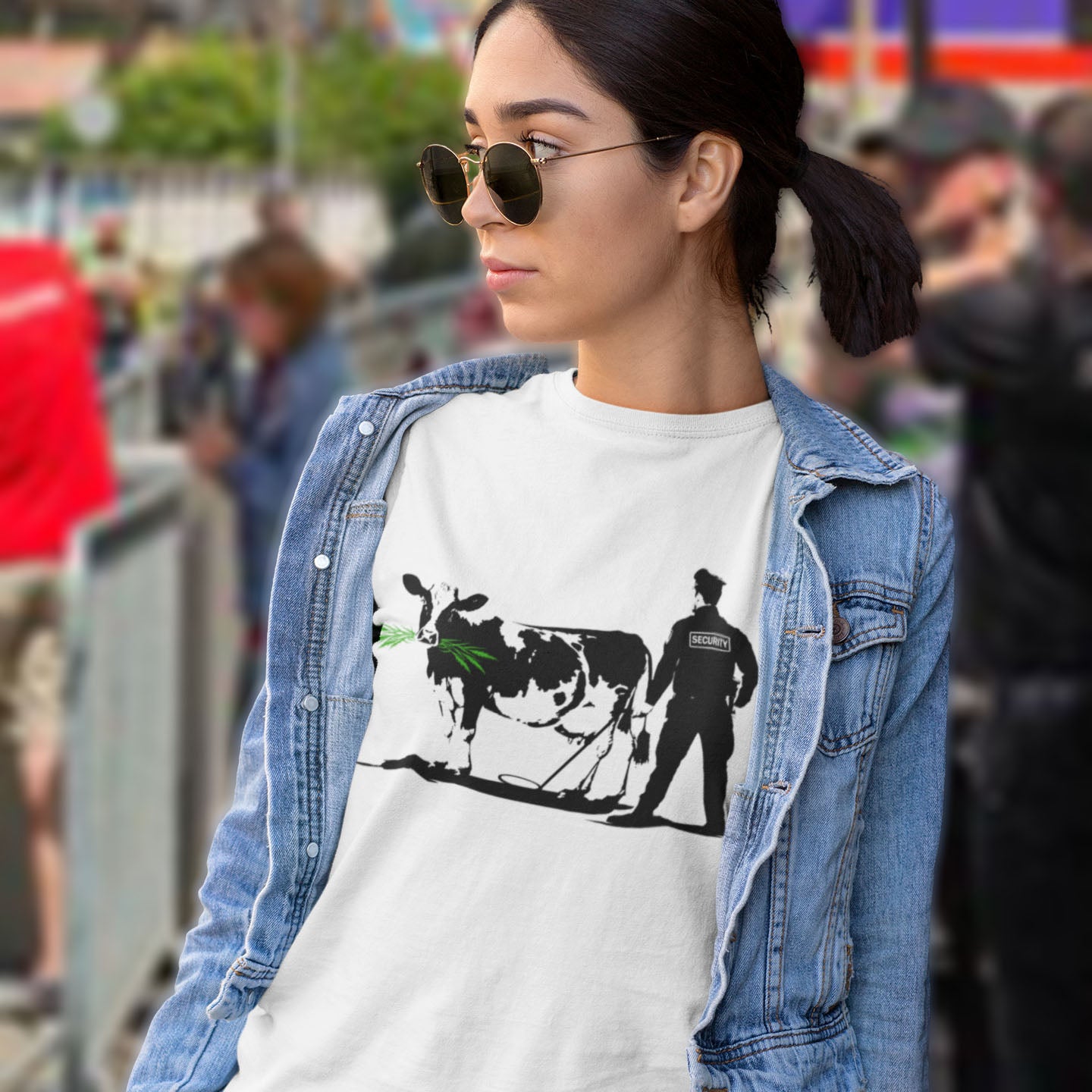 Woman wearing a white t-shirt with a graphic design of a cow and a person, standing in a crowd. A stencil graffiti t shirt showing the thin line between security and invasion of privacy, highlighted with a touch of satire and sarcasm. Made from 100% organic ring-spun cotton, this urban art t shirt is unisex, high-quality, super comfy, and best of all, eco-friendly.