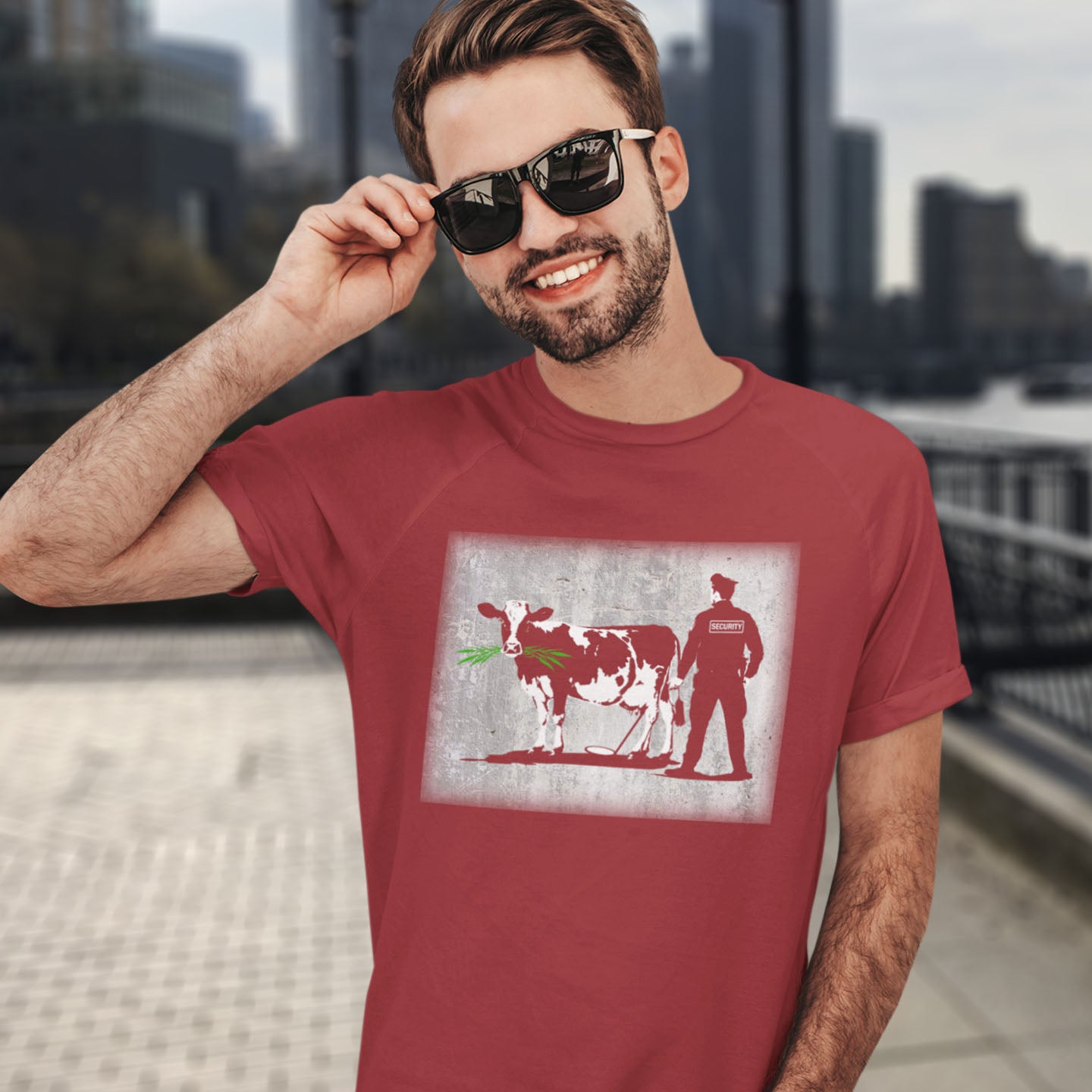 Man wearing a red t-shirt with a graphic design of a cow and person, standing in an urban setting. A graffiti stencil art t-shirt showing the thin line between security and invasion of privacy highlighted with a touch of satire and sarcasm. Made from 100% organic ring-spun cotton, this Banksy style stencil graffiti t-shirt is unisex, high-quality, super comfy, and best of all, eco-friendly.
