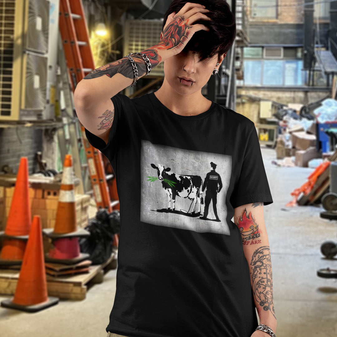 Person wearing a black t-shirt with a graphic design of a cow and a person, with tattoos on arms, in an urban setting. A stencil graffiti t shirt showing the thin line between security and invasion of privacy, highlighted with a touch of satire and sarcasm. Made from 100% organic ring-spun cotton, this urban art t shirt is unisex, high-quality, super comfy, and best of all, eco-friendly.