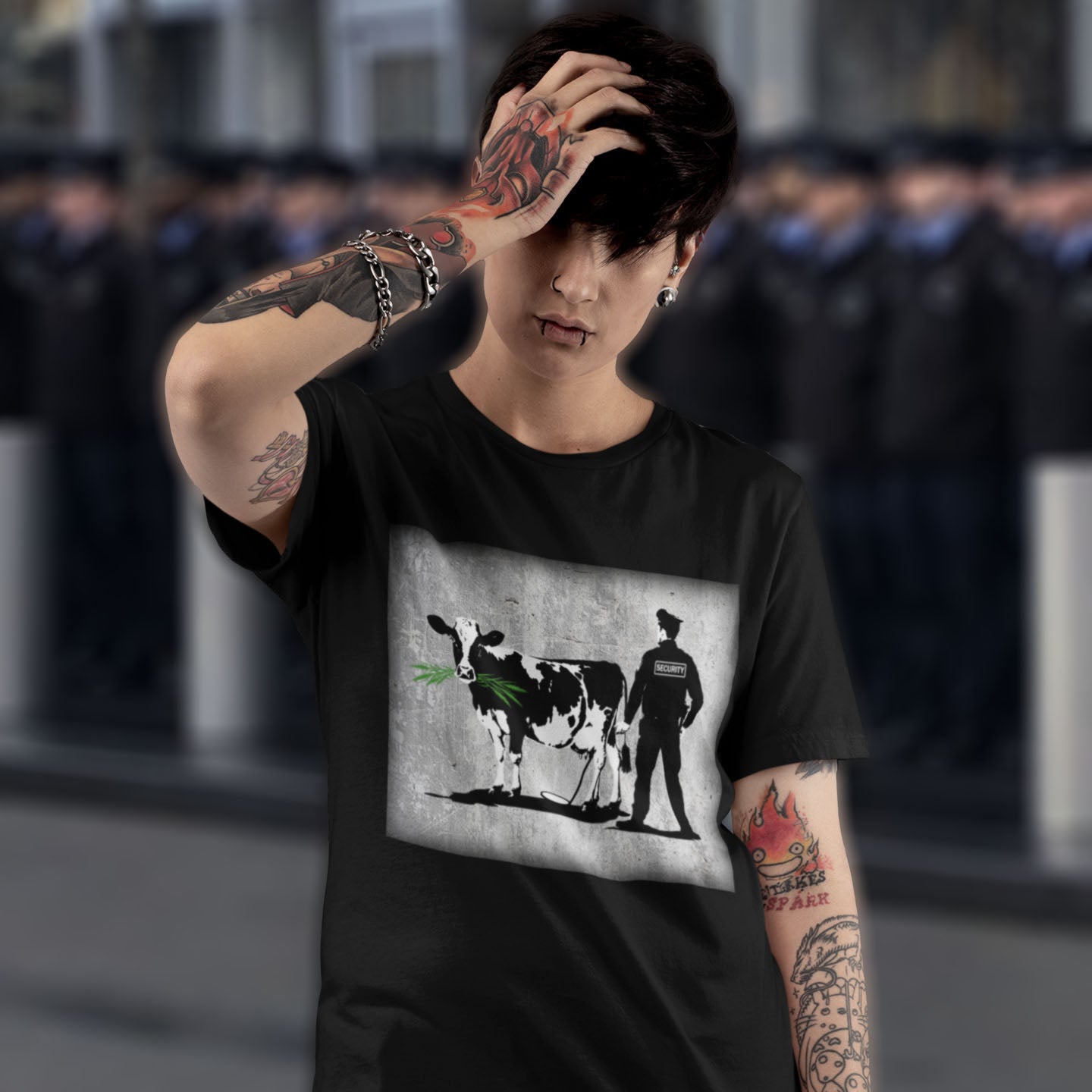 Person wearing a black t-shirt with a graphic design of a cow and a person, with tattoos on arms, in an urban setting. A graffiti stencil art t-shirt showing the thin line between security and invasion of privacy highlighted with a touch of satire and sarcasm. Made from 100% organic ring-spun cotton, this Banksy style stencil graffiti t-shirt is unisex, high-quality, super comfy, and best of all, eco-friendly.
