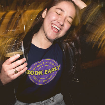 Laughing woman drinking  beer and wearing a 'book early' French navy t-shirt . A satirical and sarcastic t-shirt design highlighting the lack of spontaneity and imagination in modern life. The more efficient we make society the more we kill the essence and wonder of life. Made from 100% organic ring-spun cotton, this satirical and sarcastic t-shirt is unisex, high-quality, super comfy, and best of all, eco-friendly.