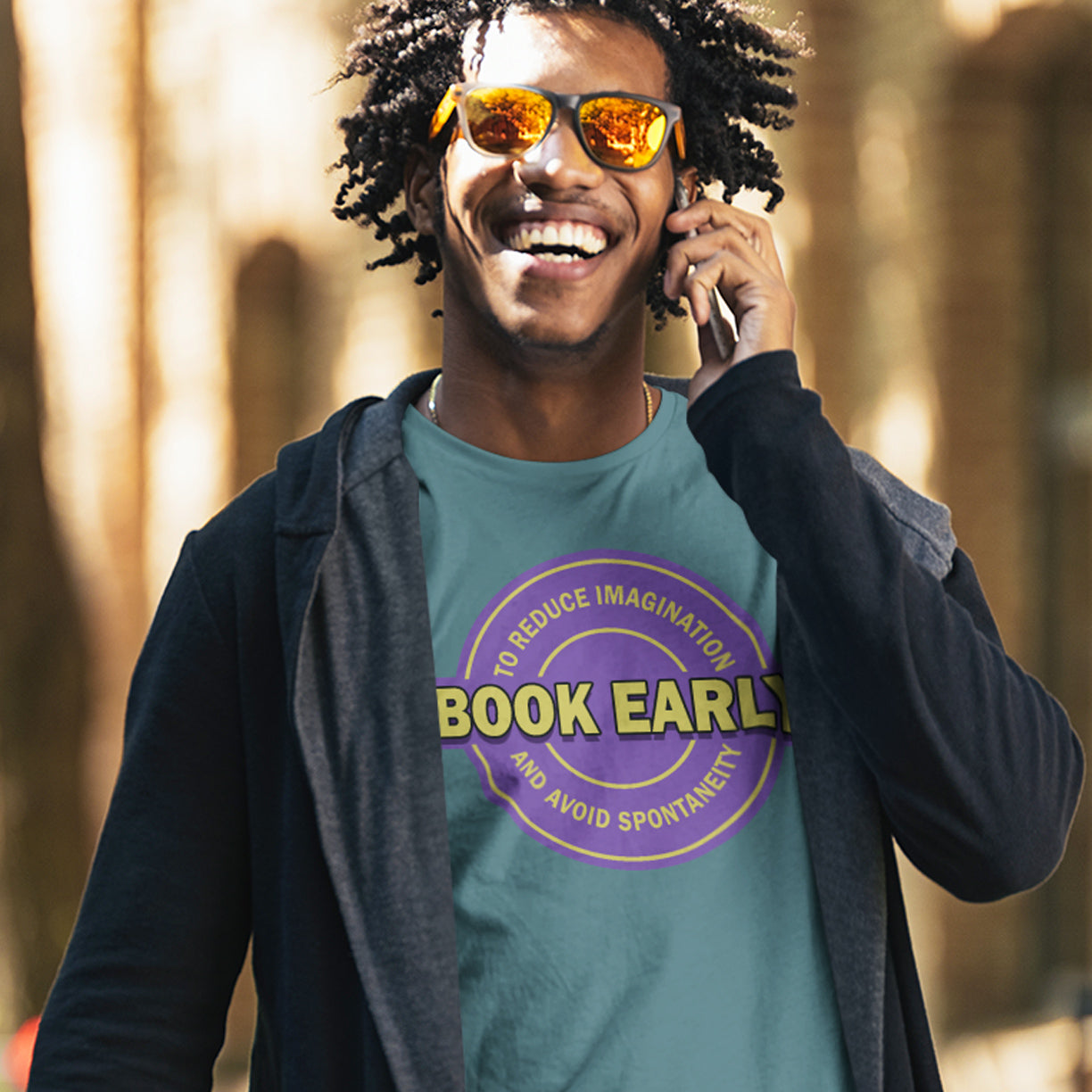 Man wearing a teal t-shirt with 'Book Early' design, talking on a phone outdoors. A satirical and sarcastic t-shirt design highlighting the lack of spontaneity and imagination in modern life. The more efficient we make society the more we kill the essence and wonder of life. Made from 100% organic ring-spun cotton, this satirical and sarcastic t-shirt is unisex, high-quality, super comfy, and best of all, eco-friendly.