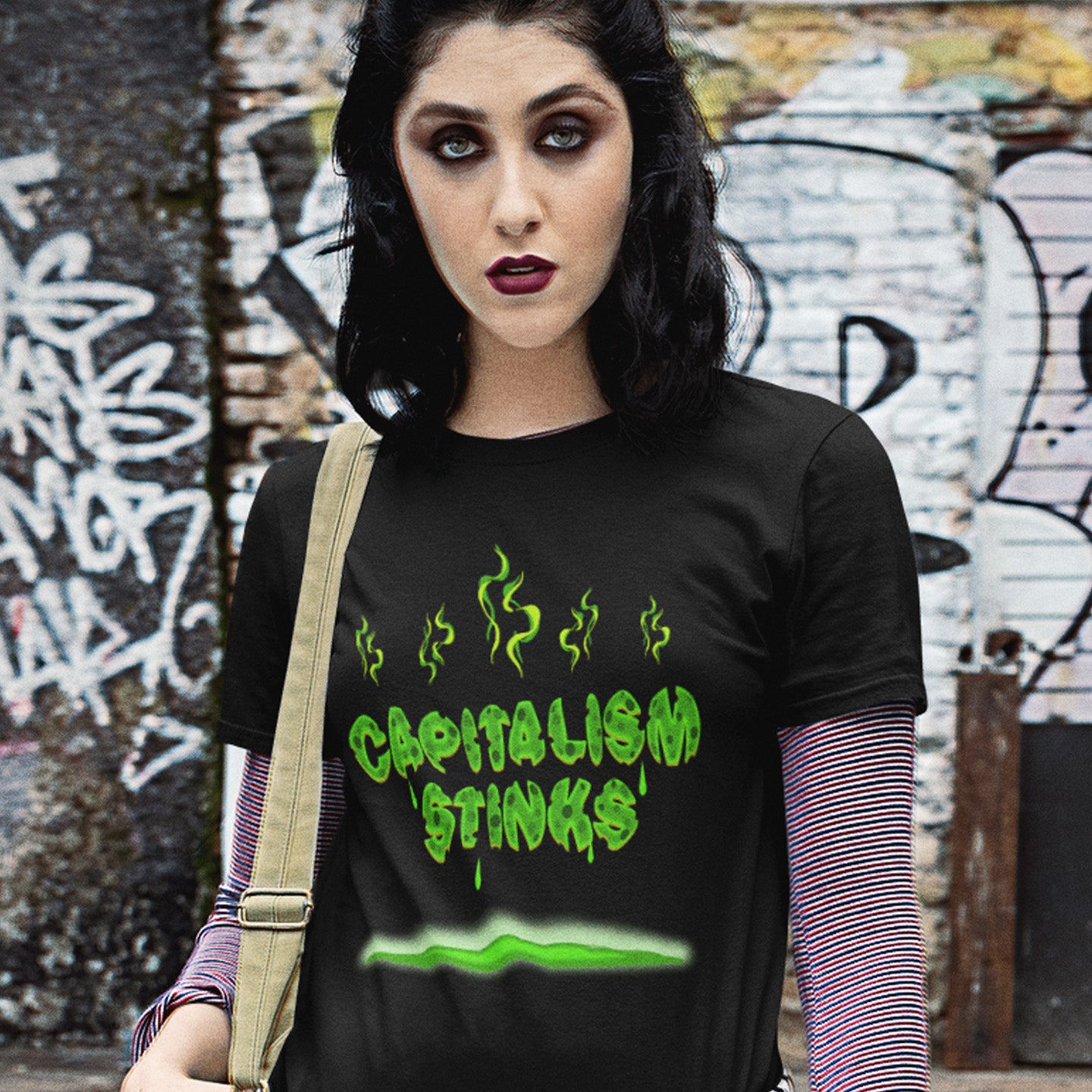 Person wearing a black t-shirt with 'CAPITALISM STINKS' text in green against a graffiti-covered wall. A funny anti capitalism t shirt exposing capitalism for what it really is. Let's face it, Capitalism has had it's day and is just rotting peoples ethics and promoting greed. Show your alternative view with this funny political t shirt. Made from 100% organic ring-spun cotton, this anti-capitalist t shirt is unisex, high-quality, super comfy, and best of all, eco-friendly.