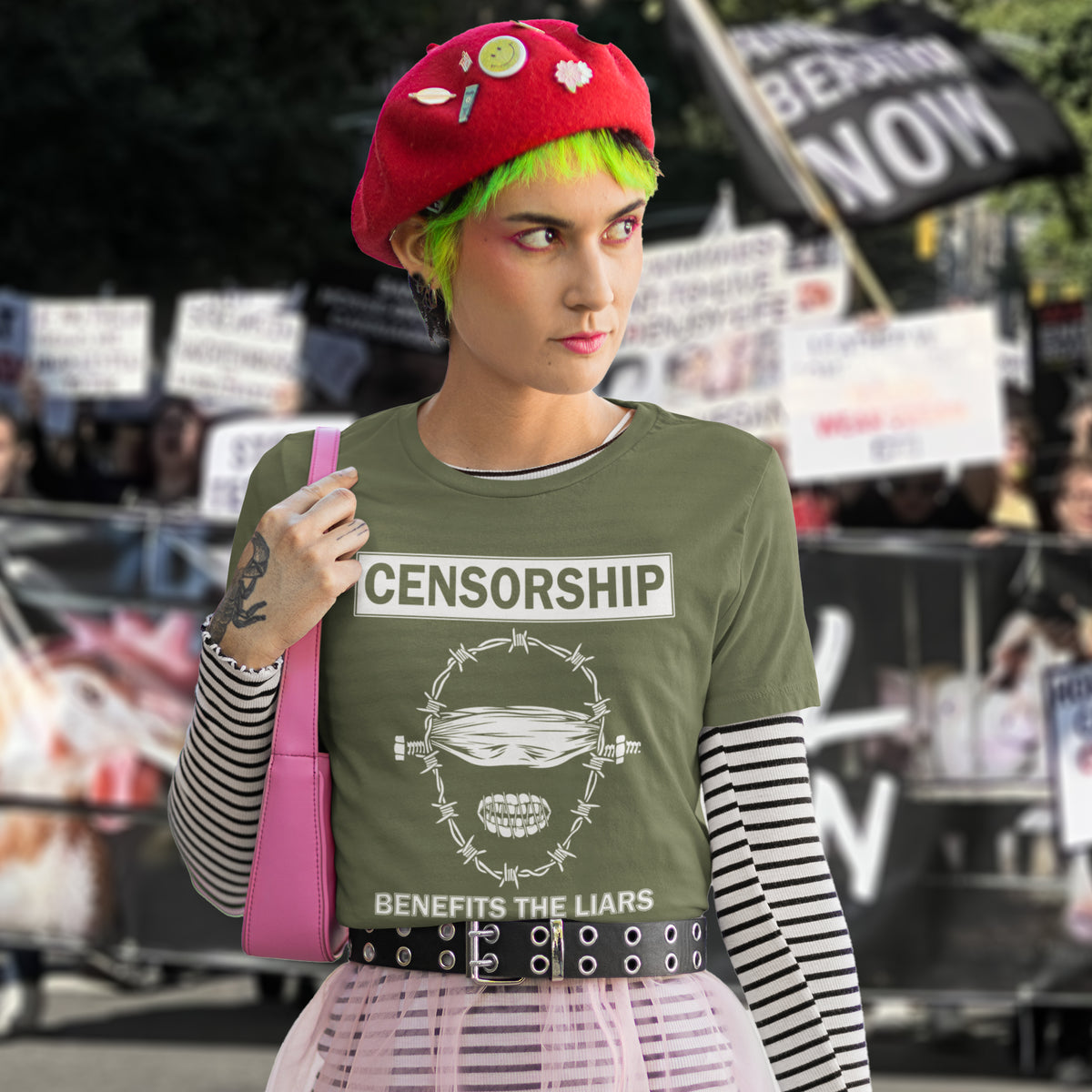 A woman with green hair and a beret, standing in front of a crowd, wearing a khaki green t-shirt with a white graphic of a face with barbed wire around it  and the text "CENSORSHIP BENEFITS THE LIARS".